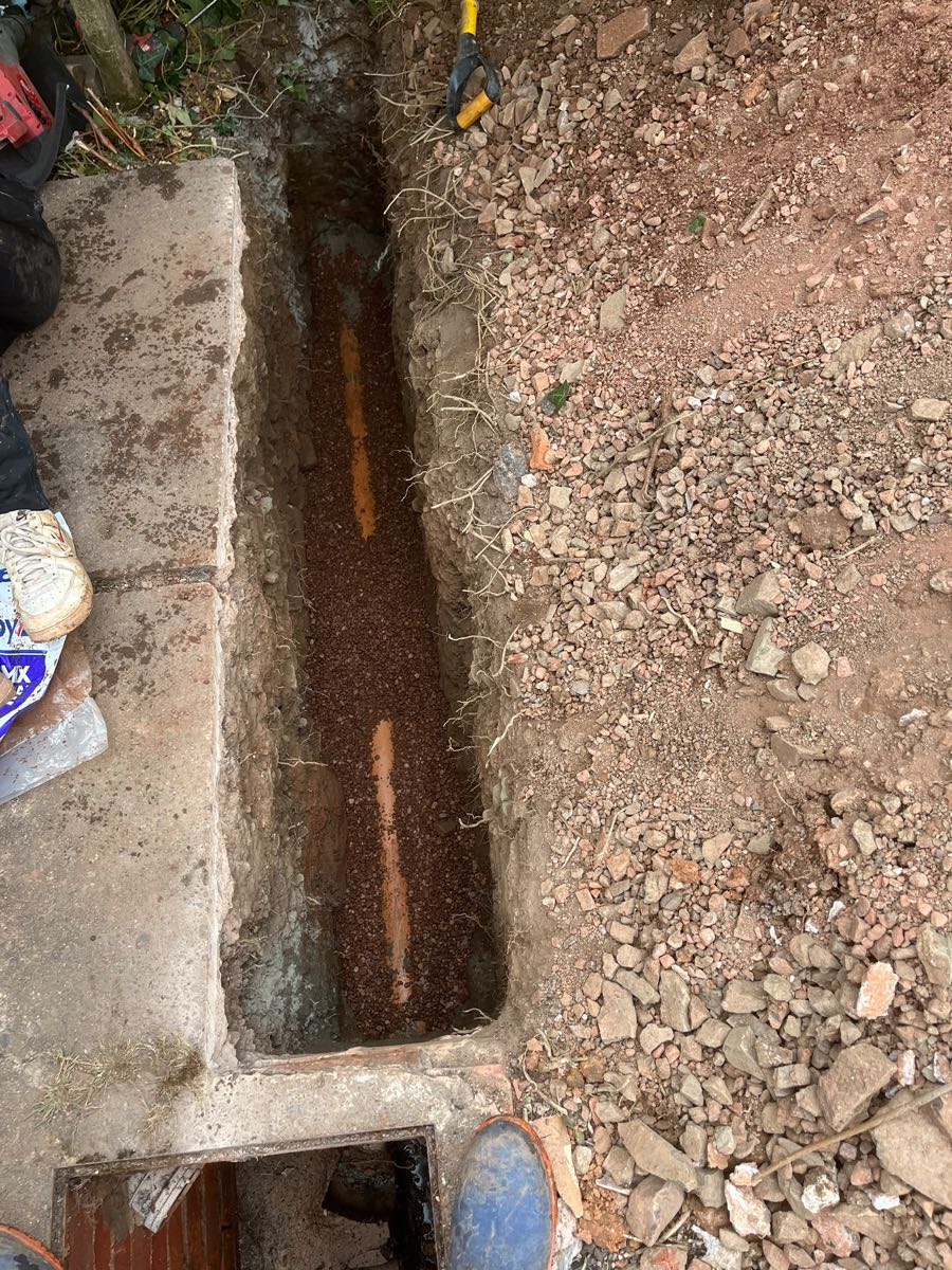 Deep narrow trench with a new drain pipe bedded in shingle running towards a chamber