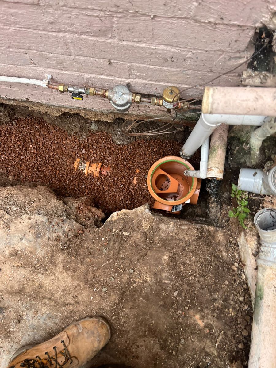 Open trench along a house wall with a new orange drain pipe bedded in shingle