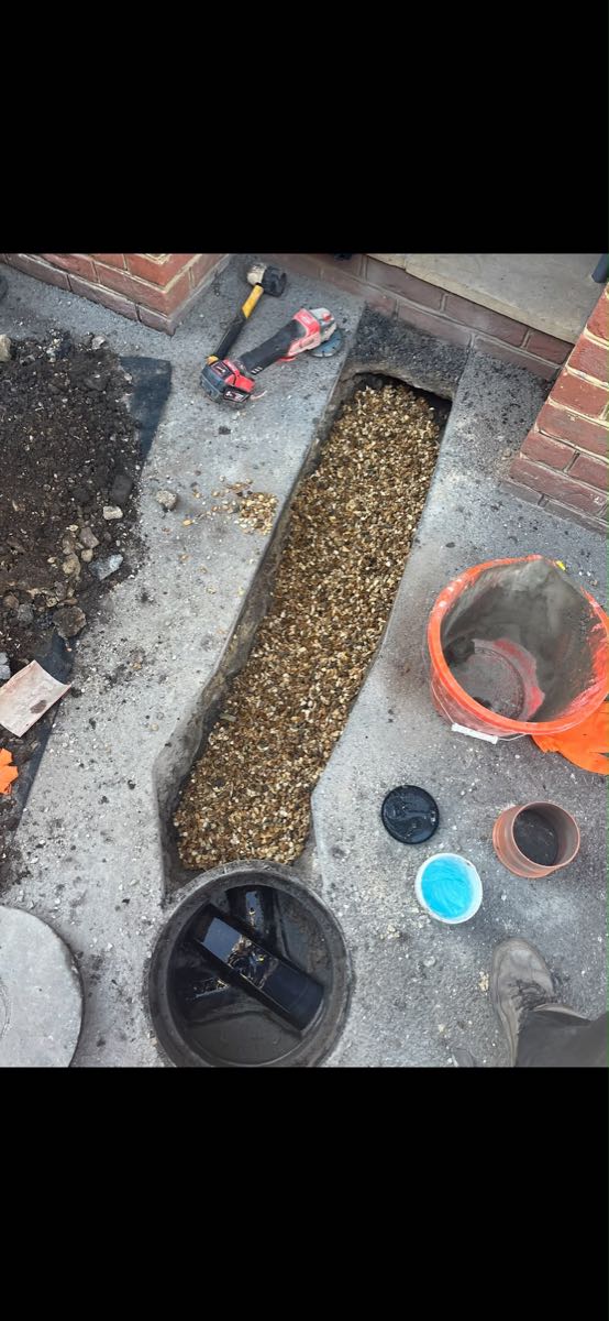 Trench cut through a concrete path filled with gravel, with the round inspection chamber open beside it